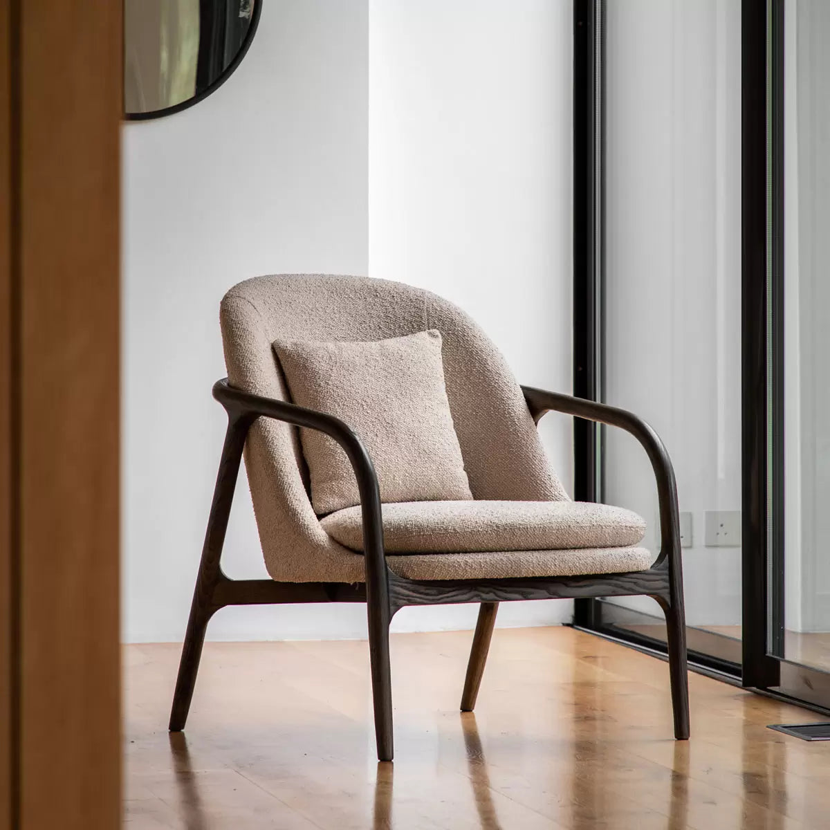 A front-three-quarter view of the Gallery Alegra taupe fabric armchair, showing its curved dark wood frame and comfortable padded taupe cushions in a sunlit room.