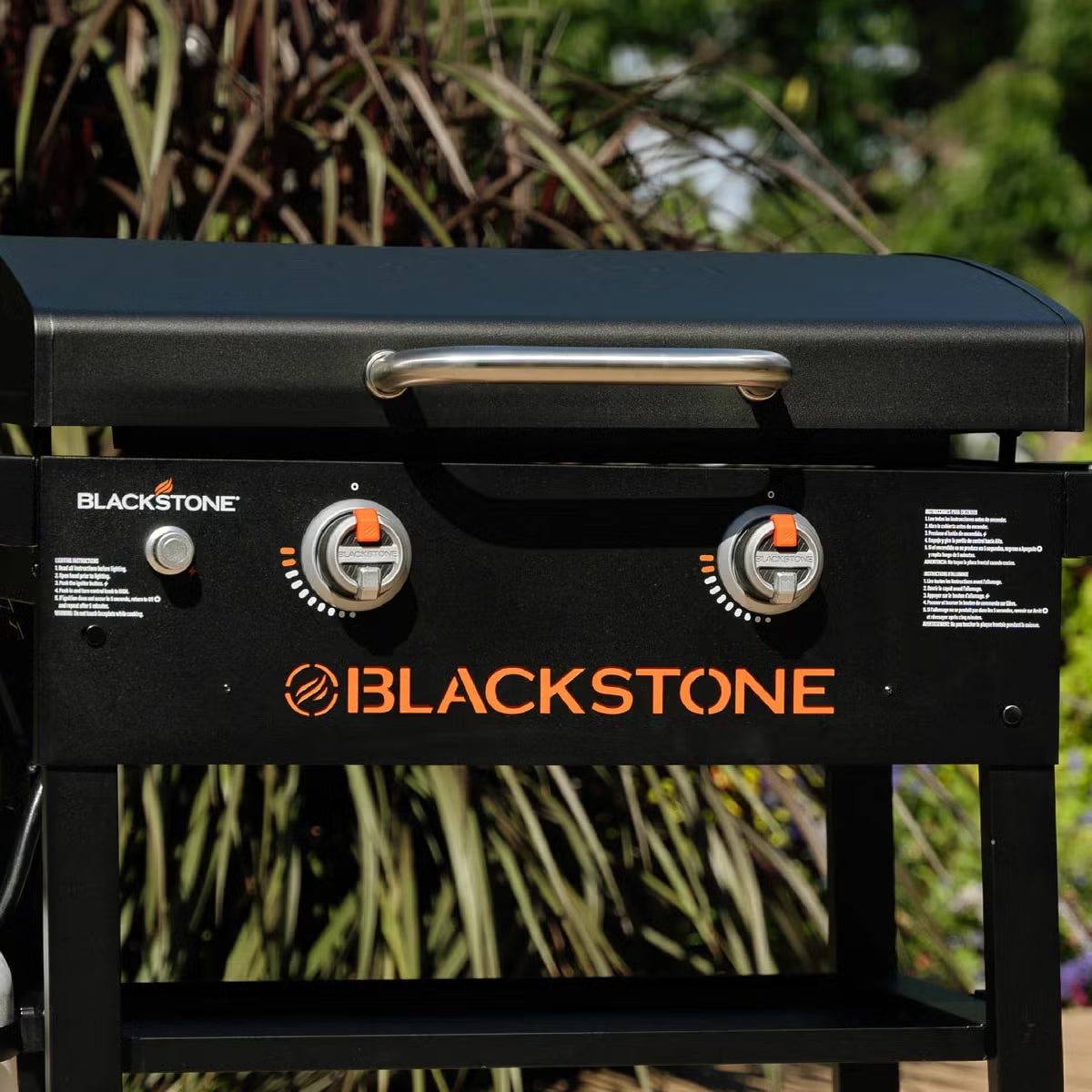 Front three-quarter view of the black Blackstone 28 inch gas griddle with hood open, showing the raw steel flat cooktop and foldable side shelves on a stone patio.