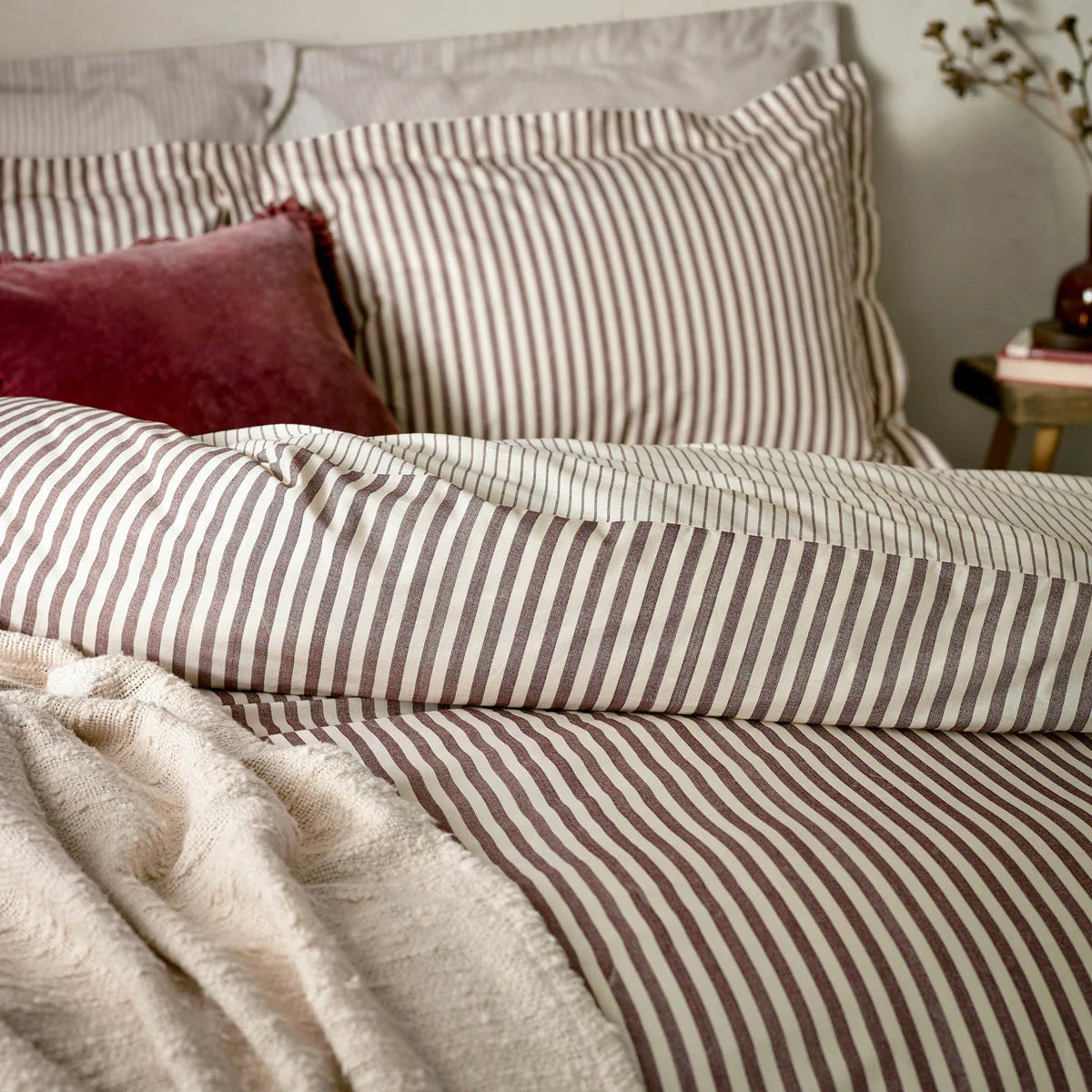 A single bed dressed in the Yard Hebden cotton burgundy striped duvet cover, showcasing the deep red and cream reversible pattern in a warm, cozy bedroom.