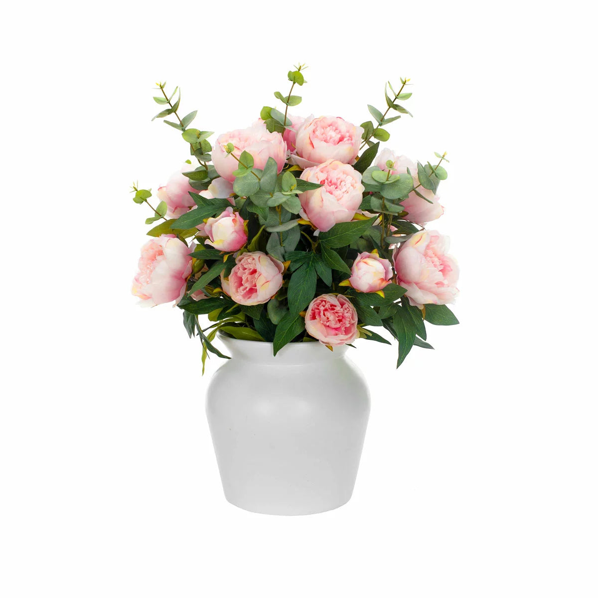 Lush artificial pink peony blooms and green eucalyptus leaves arranged inside a clear glass cylinder vase sitting on a light wooden table.