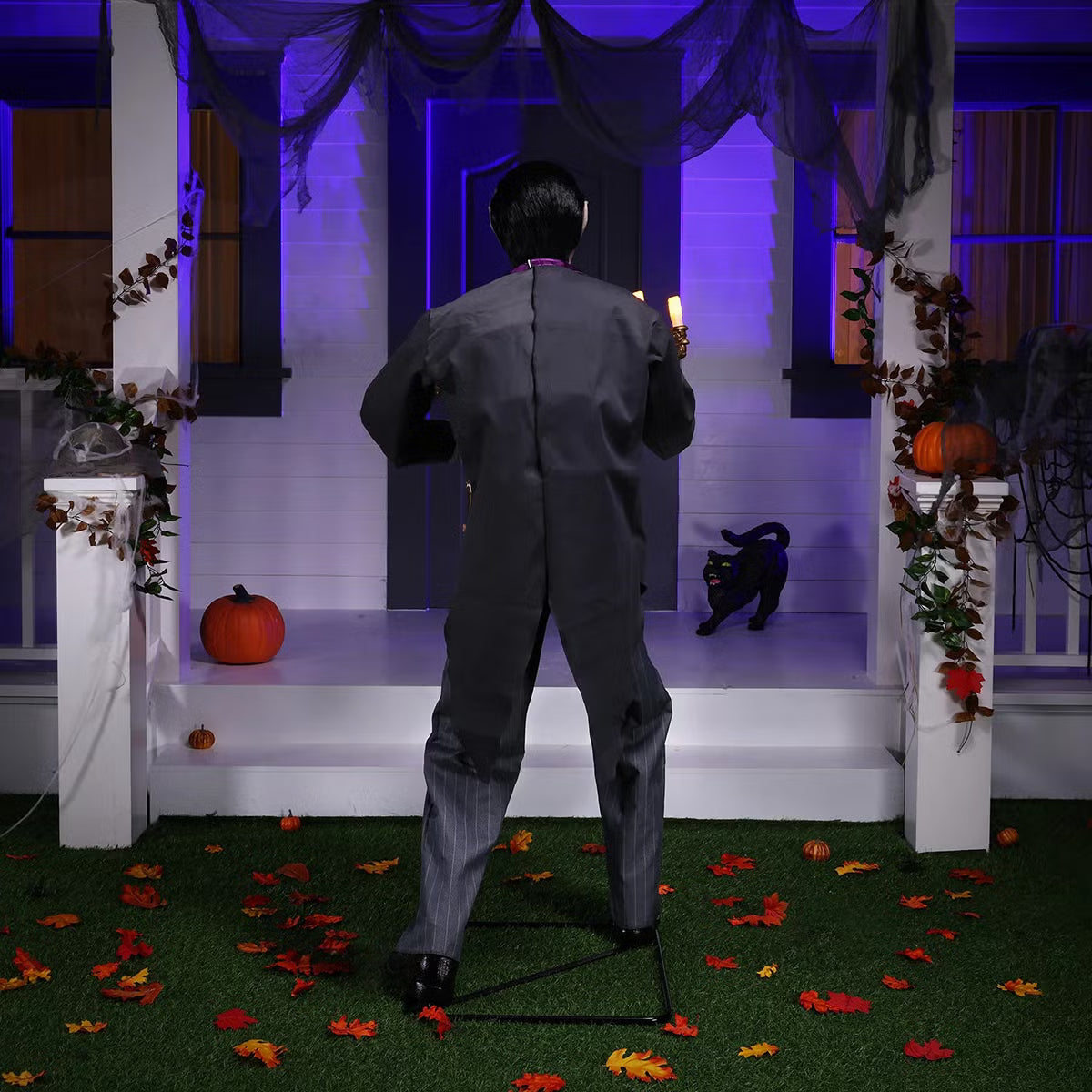 Full-length shot of a 6ft tall animated Halloween butler prop in a black suit holding an illuminated candelabra on a foggy front porch at dusk.
