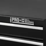 Black steel Hilka HD Pro+ 9-drawer tool chest shown from a three-quarters angle with the top lid open on dual gas struts, showcasing the aluminium drawer pulls and red interior details on a rustic wooden workbench.