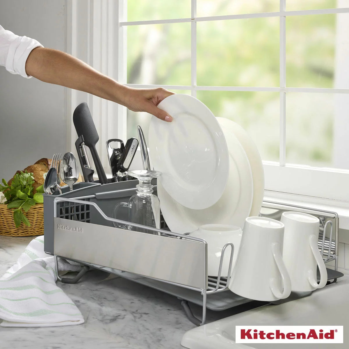 Charcoal grey KitchenAid dish rack filled with clean plates and glasses resting on a dry white quartz kitchen counter beside a stainless steel sink.