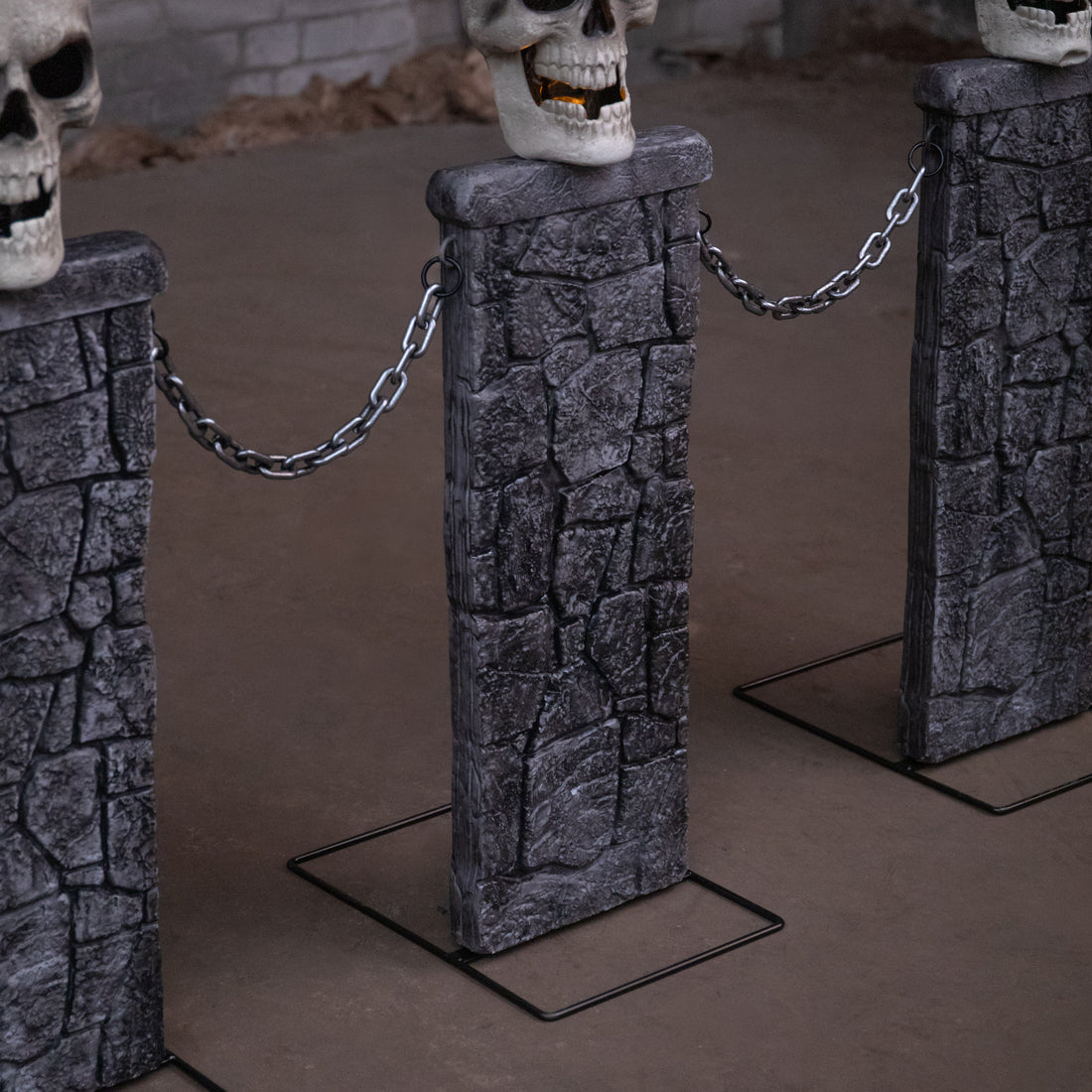 Samuel Alexander grey stone-effect Halloween skull fence with three illuminated LED pedestals and connecting chains set up on a dark lawn.