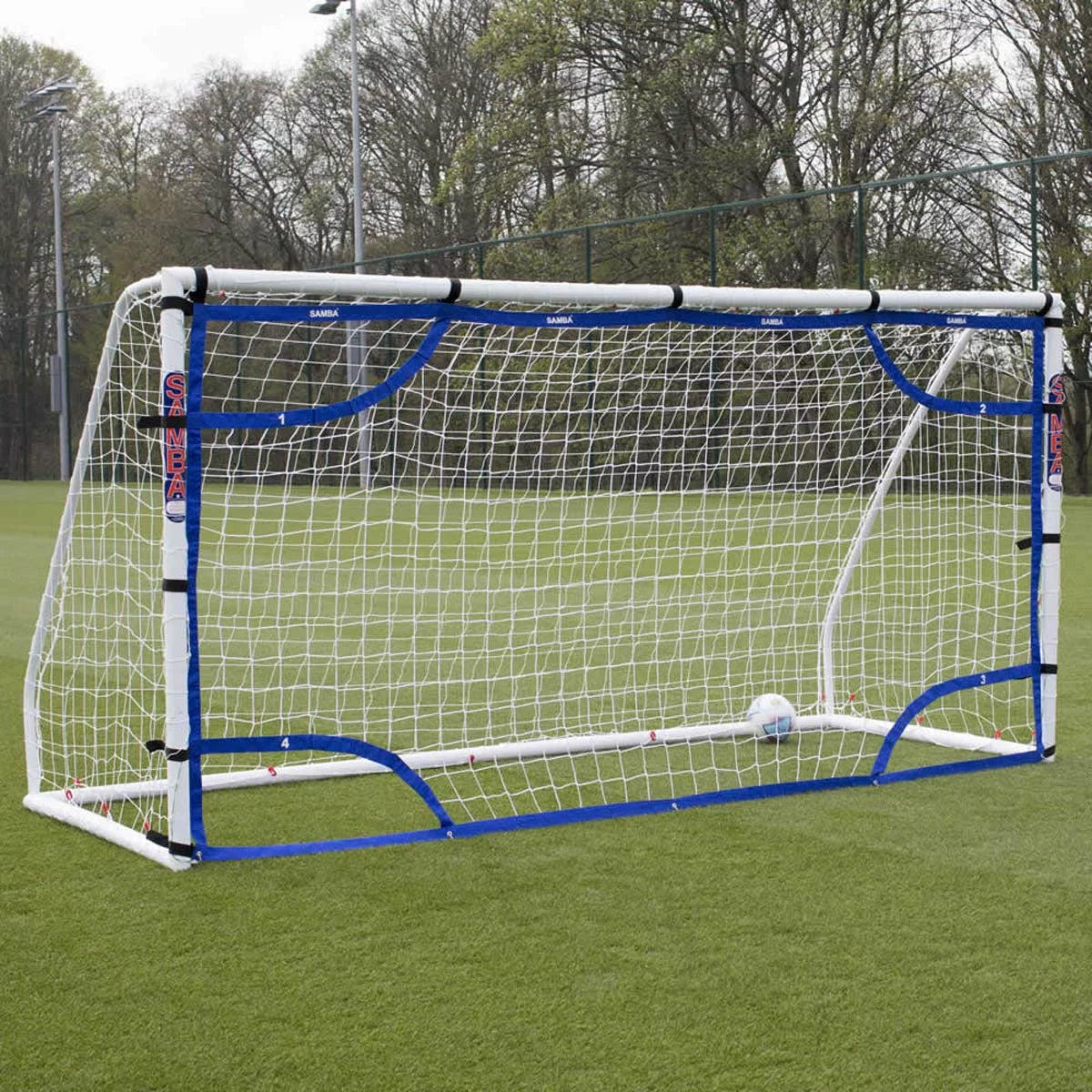 A high-visibility blue Samba 12x6 foot football target net with white numbered target zones securely attached with Velcro straps to a white goal frame on a green grass pitch.