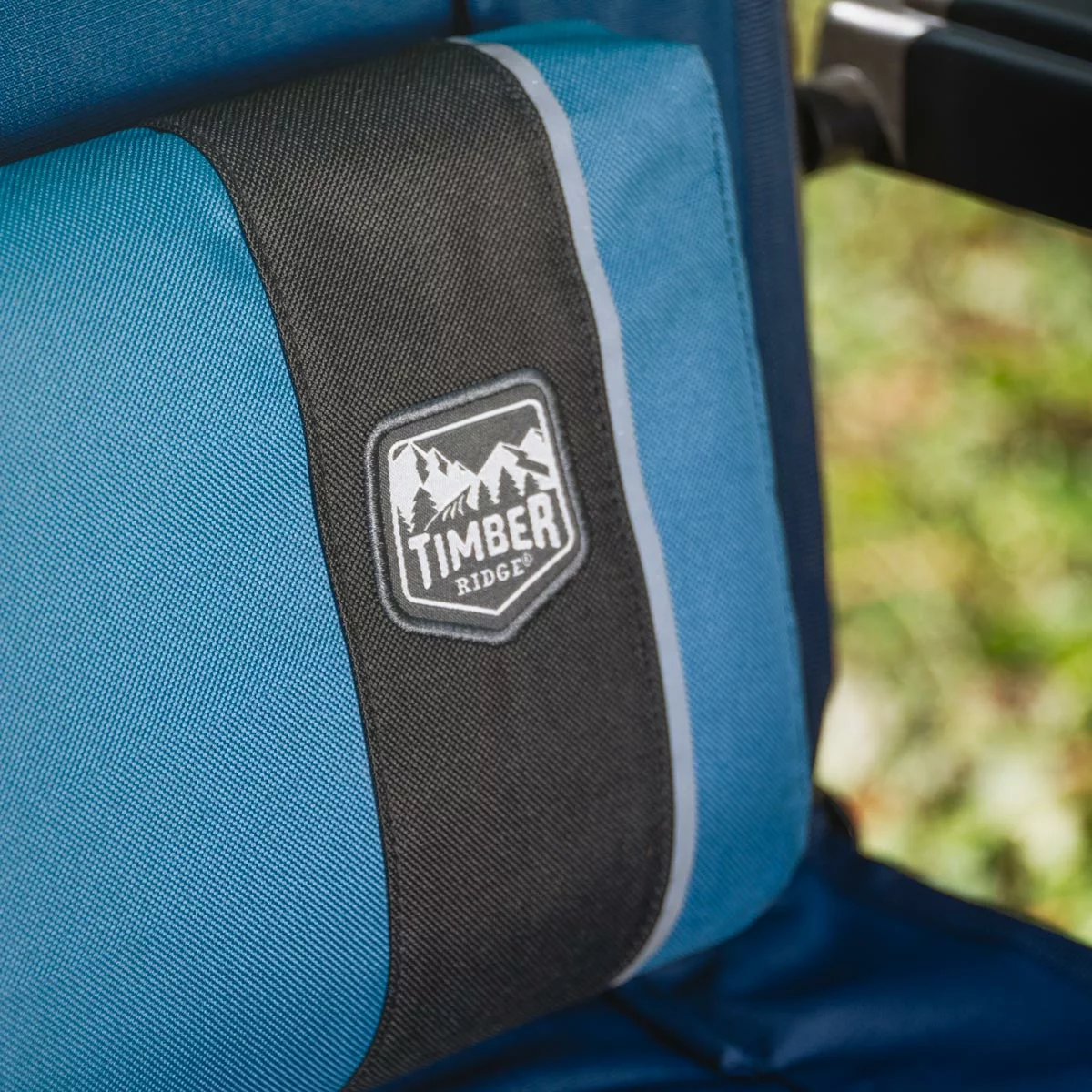 A dark padded Timber Ridge Zero Gravity Folding Lounger fully extended on a sunny outdoor patio, showcasing its open side table with a cup holder slot and the attached adjustable headrest pillow.