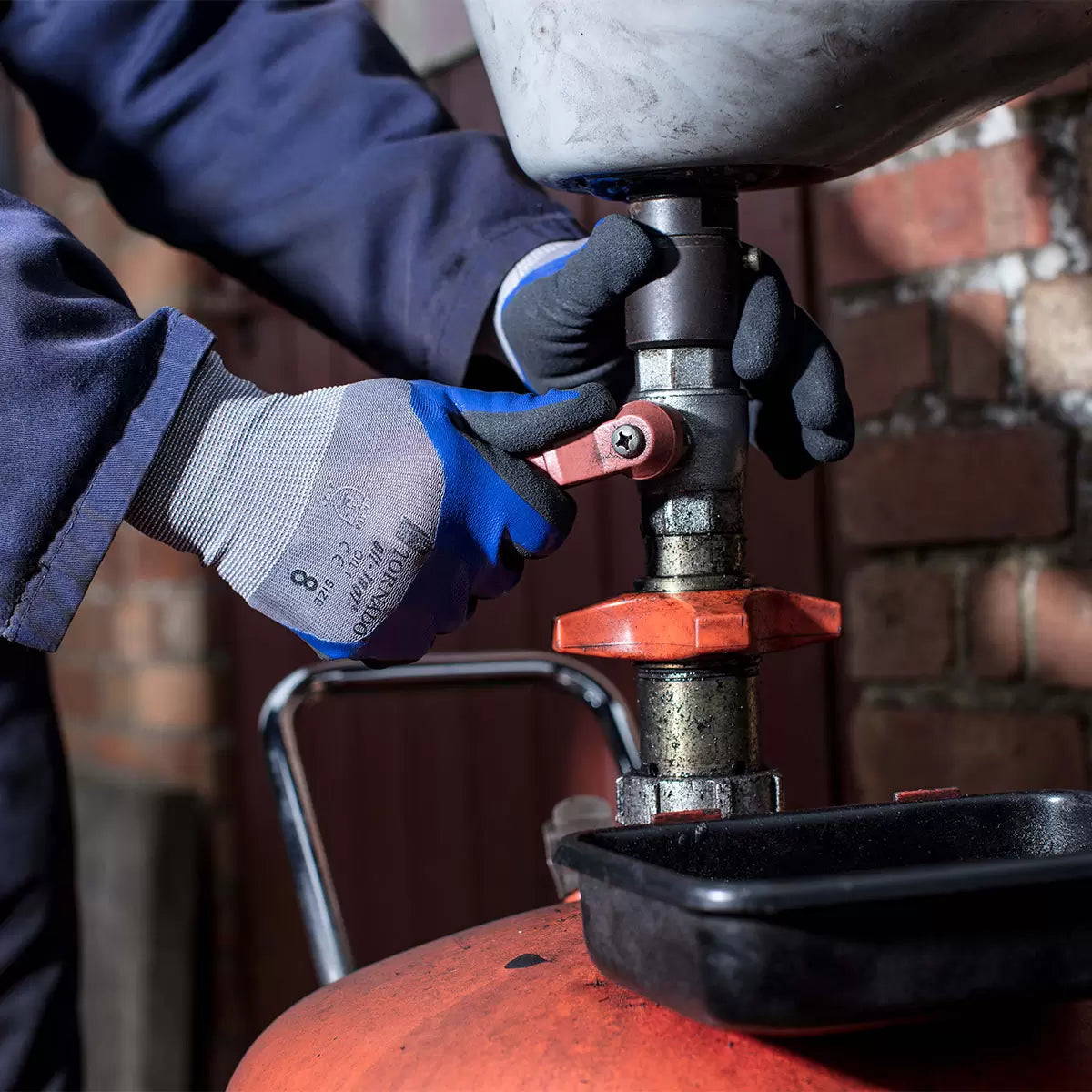 A 12-pack set of Tornado Oil-Teq 1 safety gloves showing the black and blue double-dipped nitrile palms and breathable grey back panels arranged neatly on a stainless steel workbench.