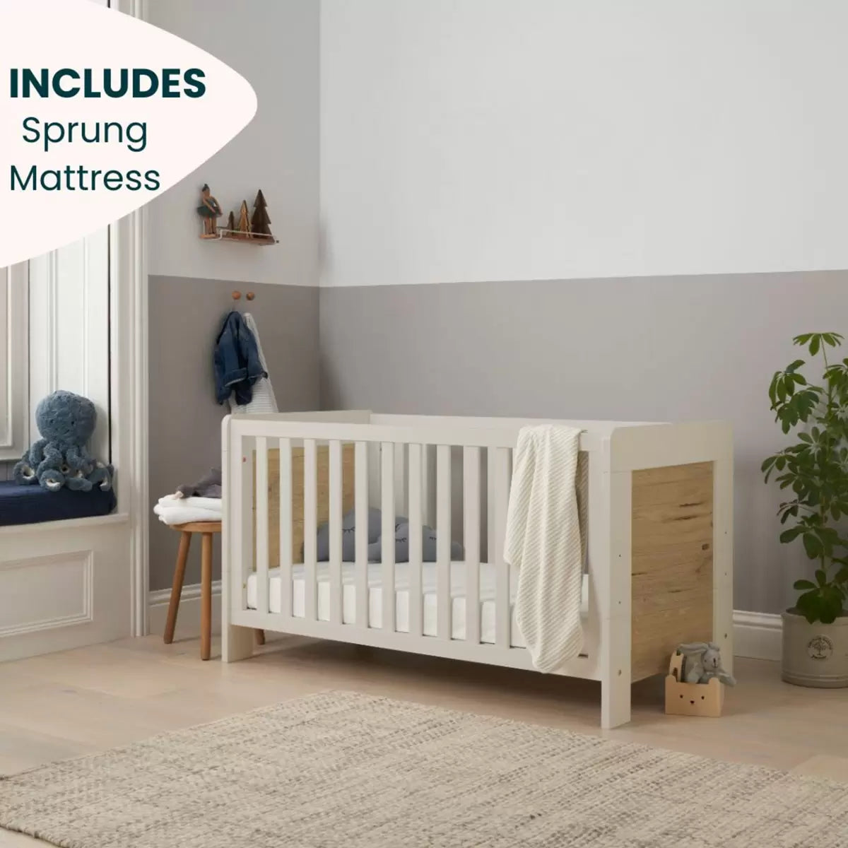 Front angle of the Tutti Bambini Alba Cot Bed in White Sand and Distressed Oak finish showing the included sprung mattress inside a brightly lit nursery setting.