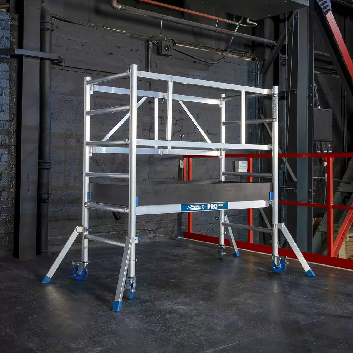 A silver aluminium Werner PROPLUS folding scaffold tower base pack with black swivel castors, a platform deck featuring an integrated trapdoor, and protective wooden toe boards set against a plain backdrop.