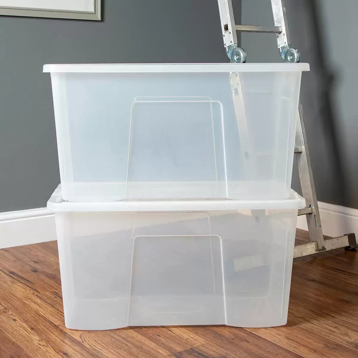 Three stacked Wham 110 litre clear plastic storage boxes with blue clip-on lids filled with blankets and clothes inside a brightly lit home closet.