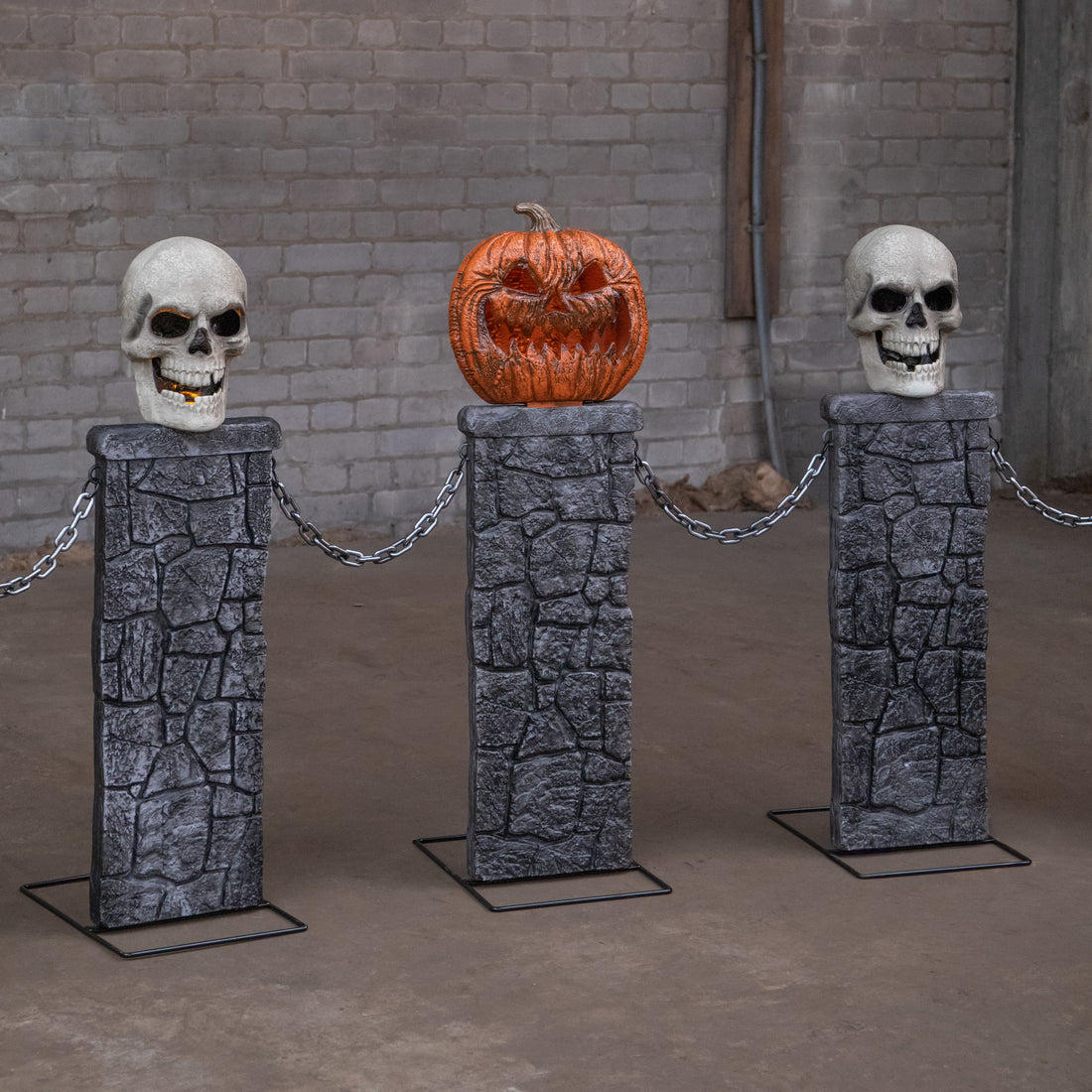 Samuel Alexander 2.4m battery powered Halloween fence featuring stone-effect tombstone pedestals with lit skull and pumpkin tops linked by black chains on a dark lawn.
