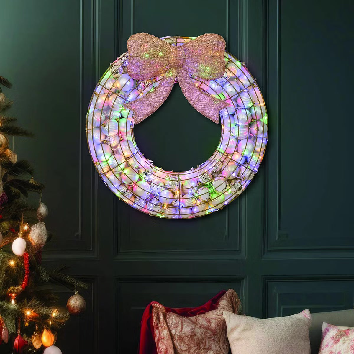 Frontal view of a 24-inch red indoor holiday wreath made of glossy and matte ornaments glowing with warm white LED lights over a dark brick mantelpiece.