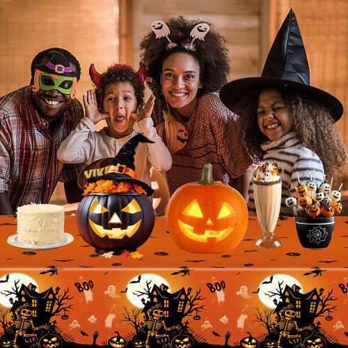 Flat lay of two orange rectangular plastic Halloween tablecloths featuring black prints of spooky castles, bats, spiders, and pumpkins spread over a long dinner table.