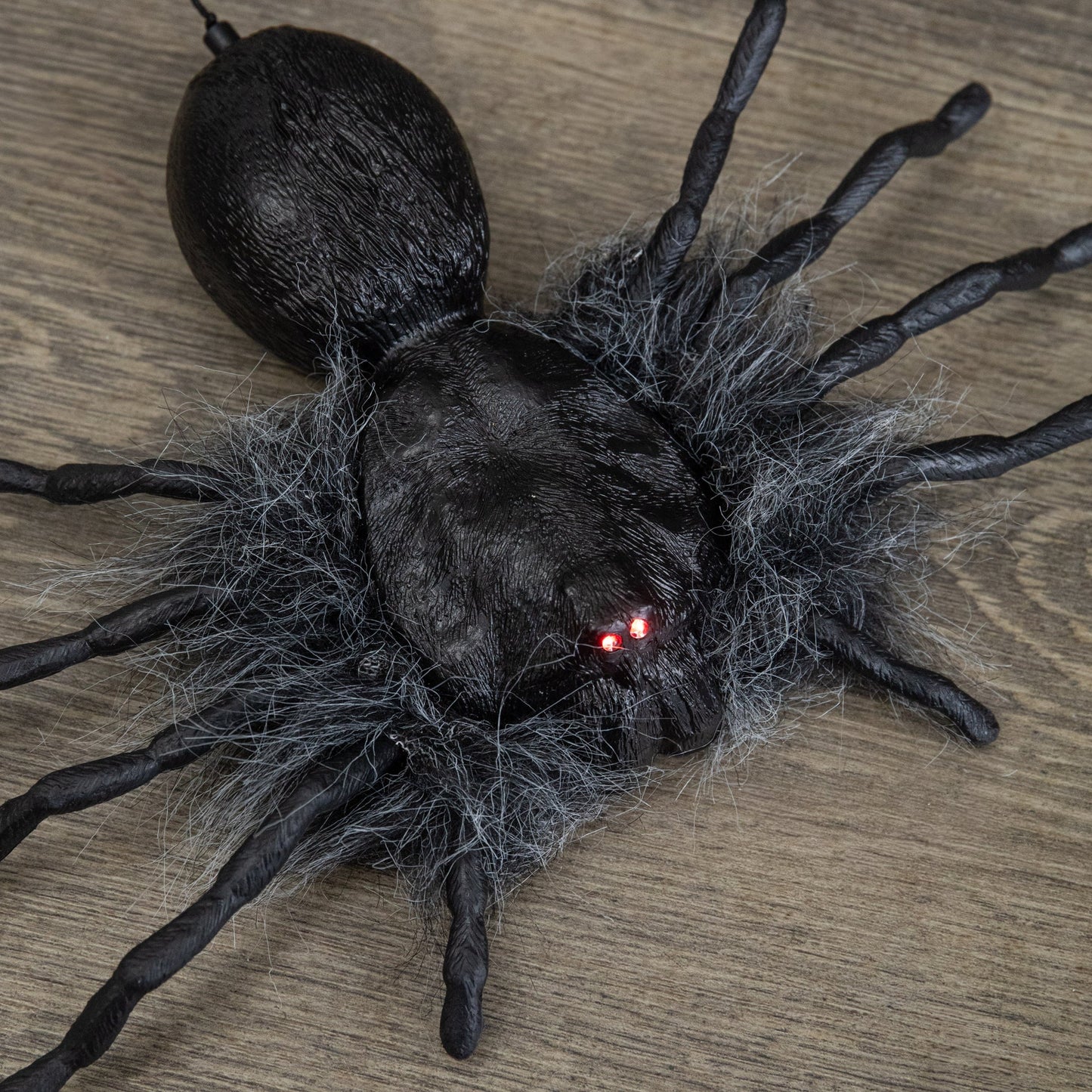 Black faux-fur animatronic spider with glowing red eyes hanging from a suspended string against a dark background.