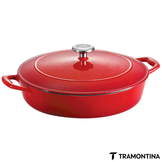 Vibrant red and blue enamelled cast iron shallow casserole dish by Tramontina with cream interior and stainless steel lid knob on a rustic wooden dining table.