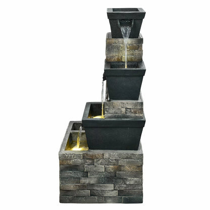 A 34.5-inch tiered brick-effect fountain glowing with warm LED lights on a stone patio, surrounded by green foliage at dusk