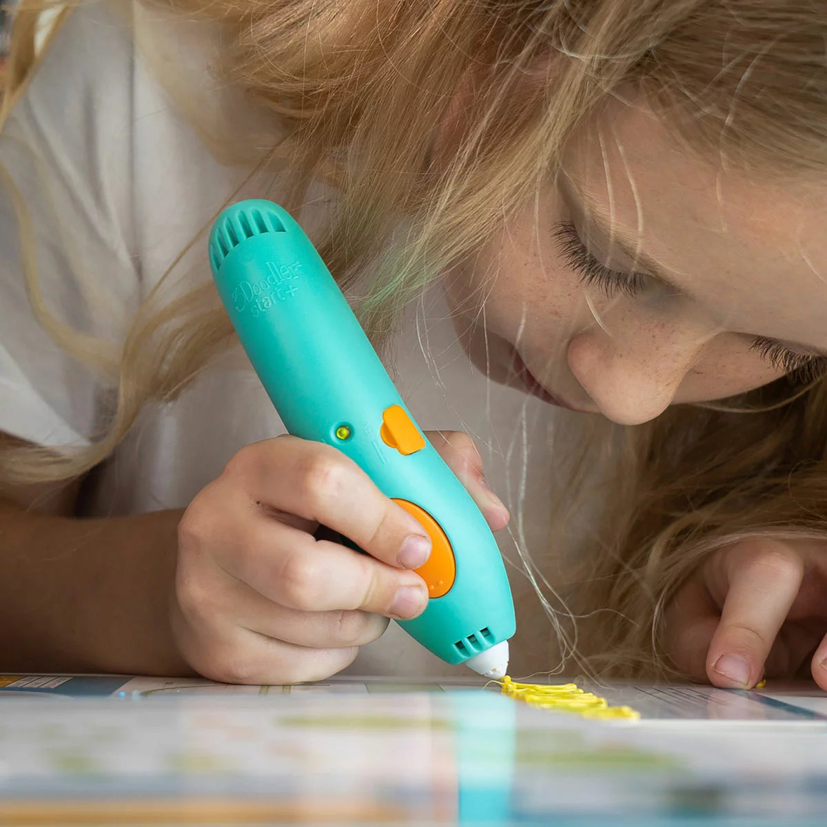 Blue 3Doodler Start+ 3D pen resting on a clear DoodlePad next to colorful eco-plastic strands, silicone molds, and an activity guide on a white play desk.