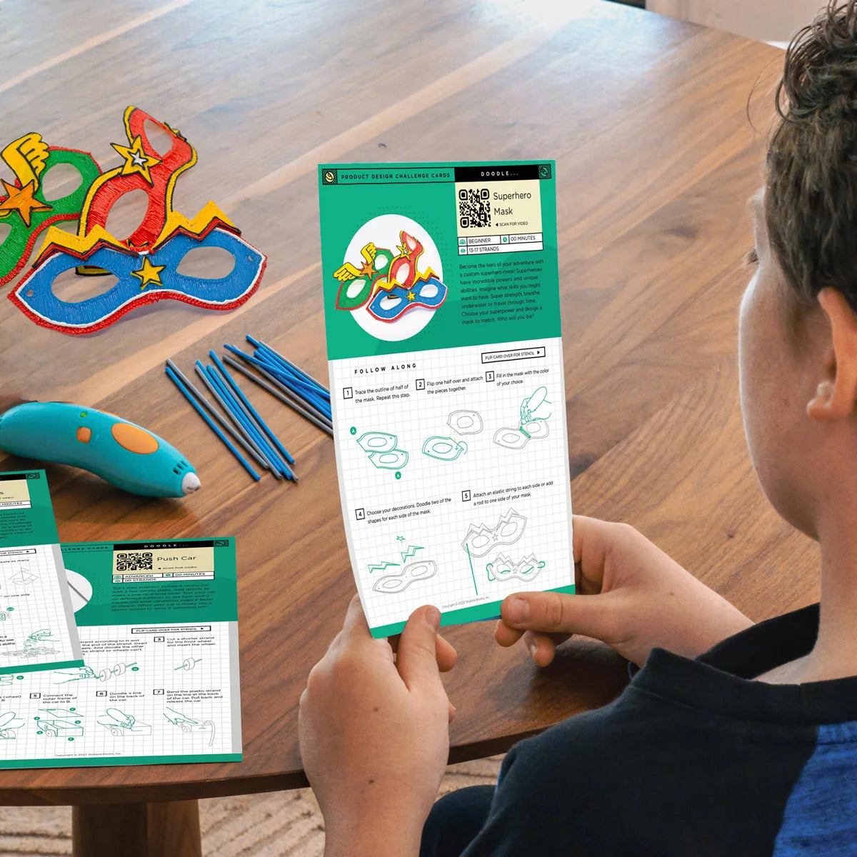 Blue 3Doodler Start+ 3D pen resting on a clear DoodlePad next to colorful eco-plastic strands, silicone molds, and an activity guide on a white play desk.