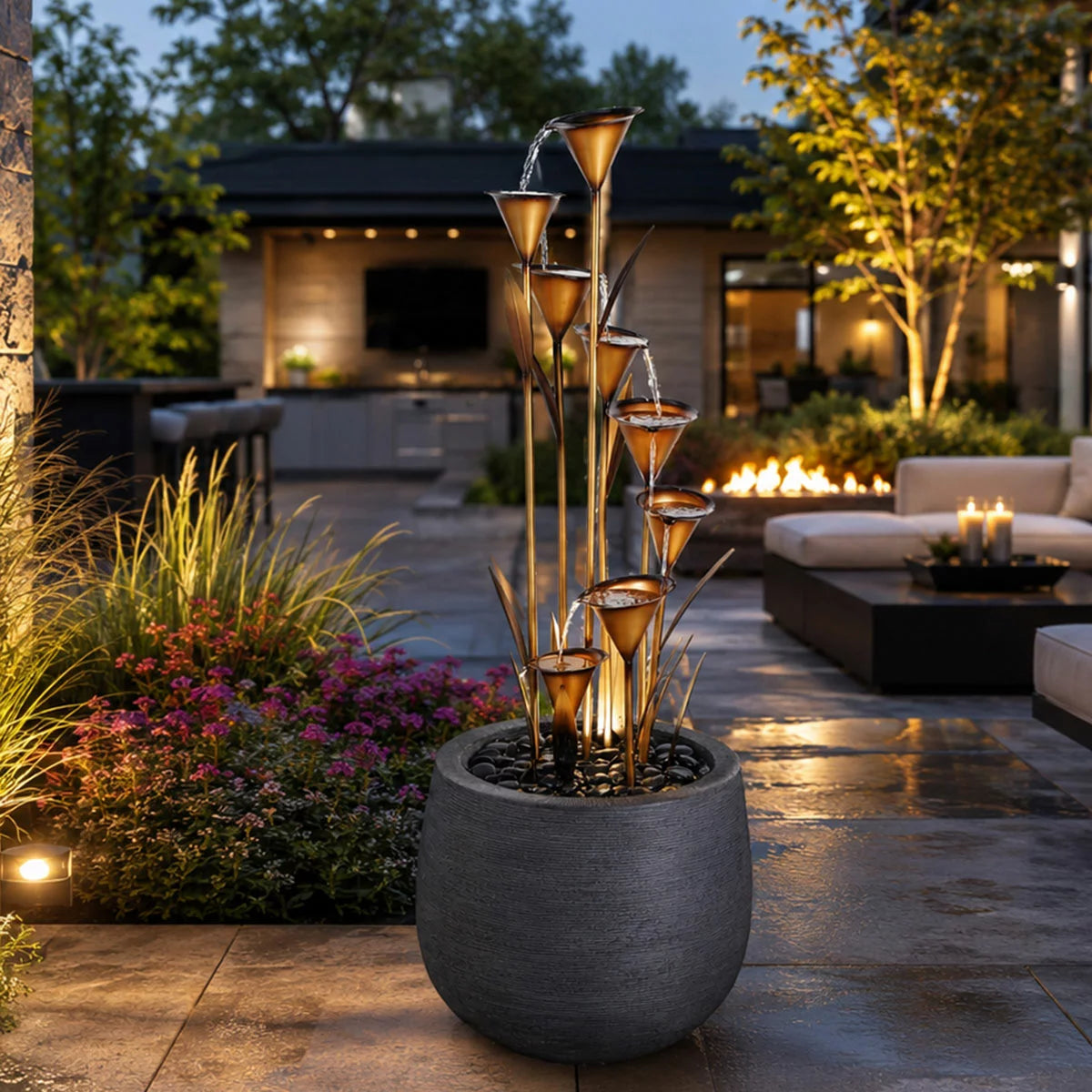A 139cm copper-finished calla lily fountain cascading water into a black river rock basin, set against a lush green garden background on a sunny day.