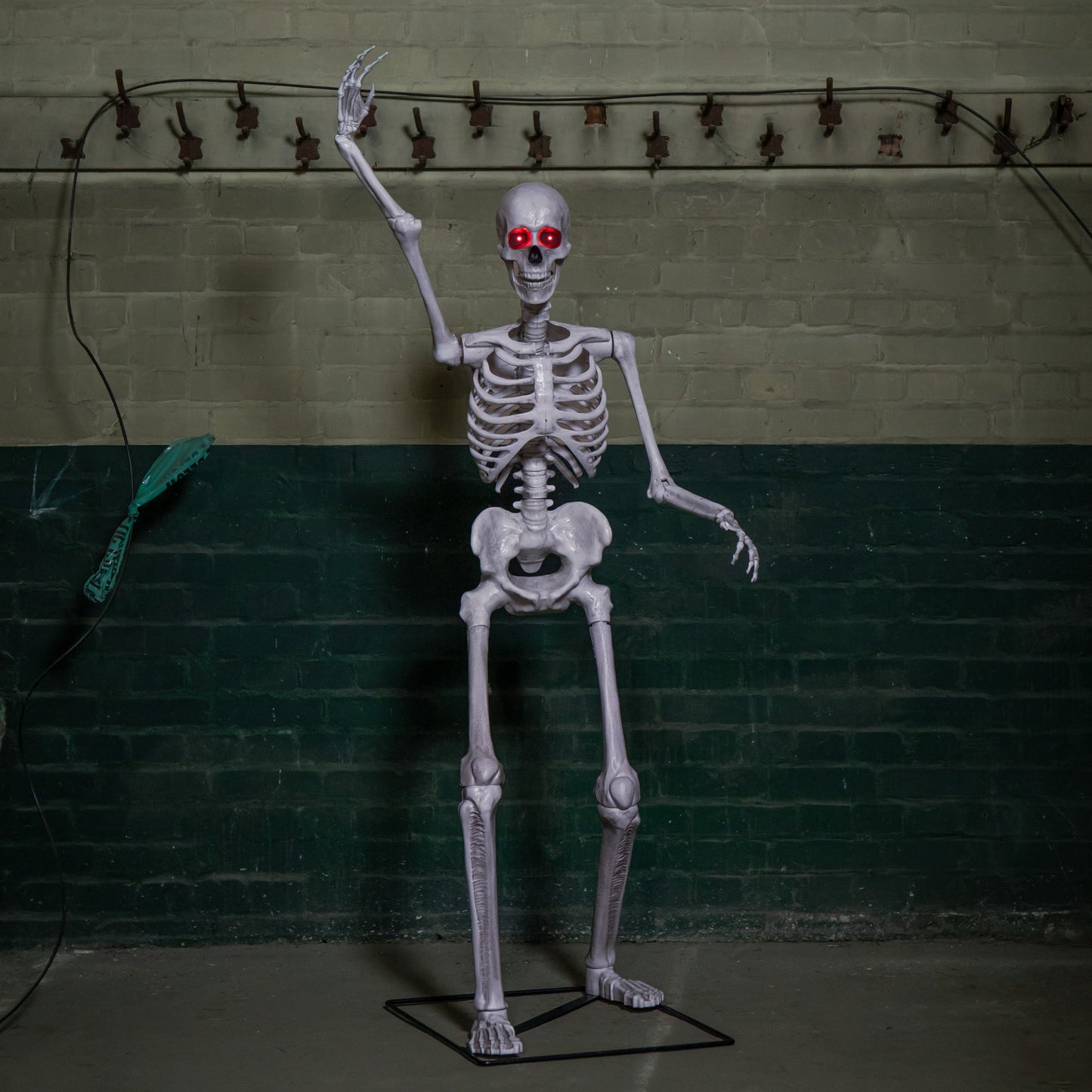 Front view of a grey 5ft full body Halloween skeleton with glowing red LED eyes standing upright on a black metal base on a porch.