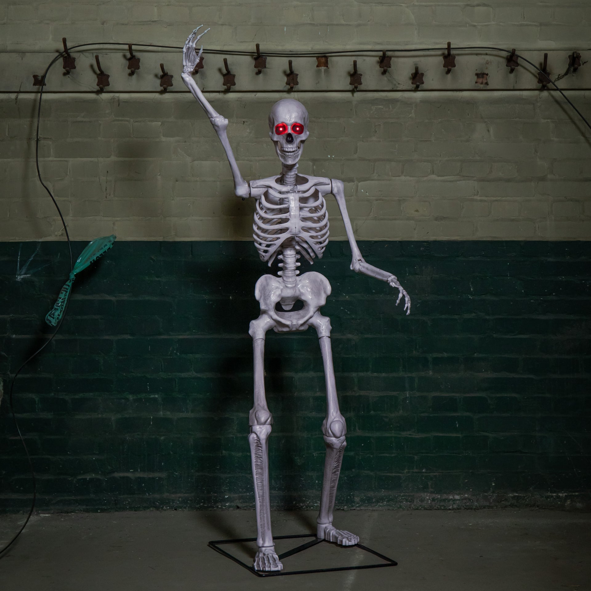 Front view of a grey 5ft full body Halloween skeleton with glowing red LED eyes standing upright on a black metal base on a porch.