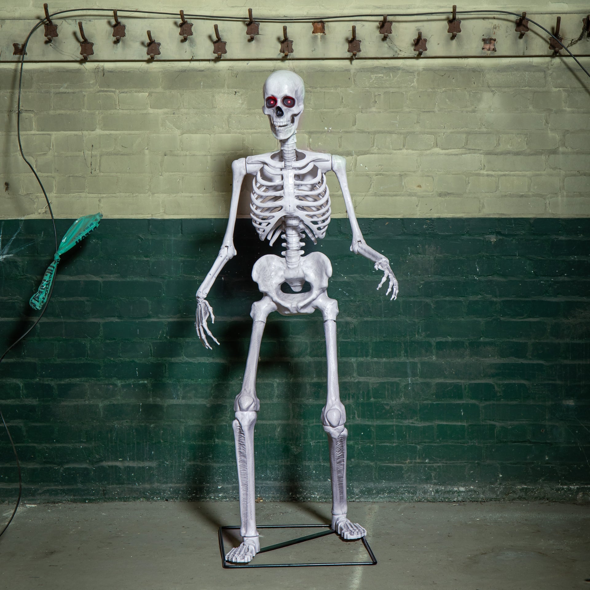 Front view of a grey 5ft full body Halloween skeleton with glowing red LED eyes standing upright on a black metal base on a porch.