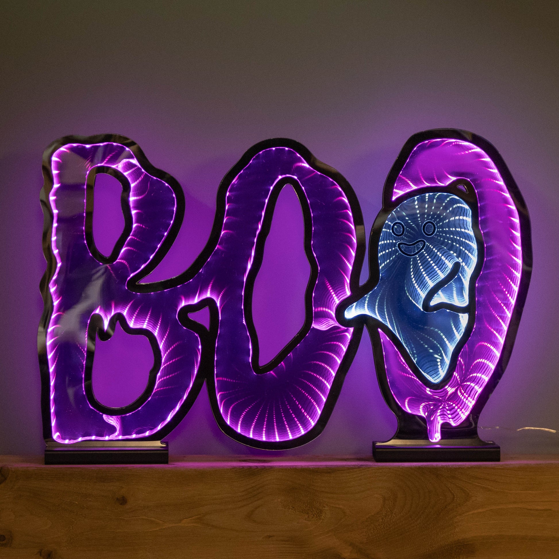 Samuel Alexander 60cm illuminated BOO infinity mirror sign with glowing pink and white LEDs mounted on a wooden base displayed on a fireplace mantel.