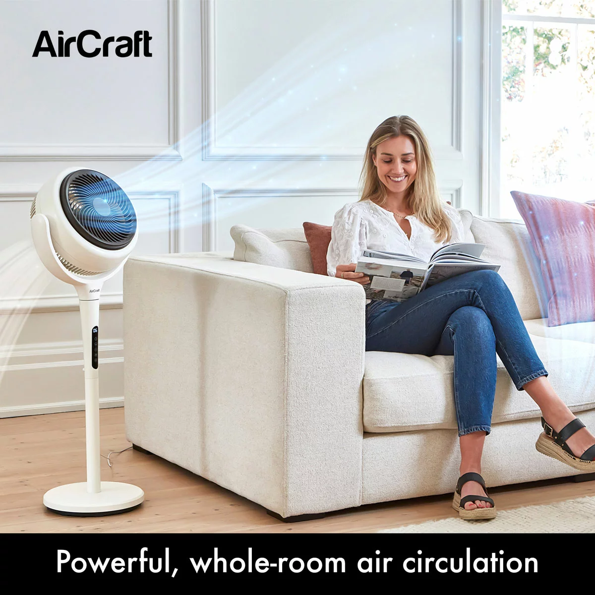 Convertible stone white AirCraft LUME 2-in-1 air circulator fan with illuminated rear backlight standing on a wooden floor beside a lounge sofa.