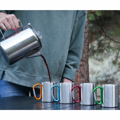 Stainless steel 9-cup Alpine Mountain Gear coffee percolator with glass top knob heating over a campfire next to four carabiner-handle stainless steel mugs on a wooden campsite table.