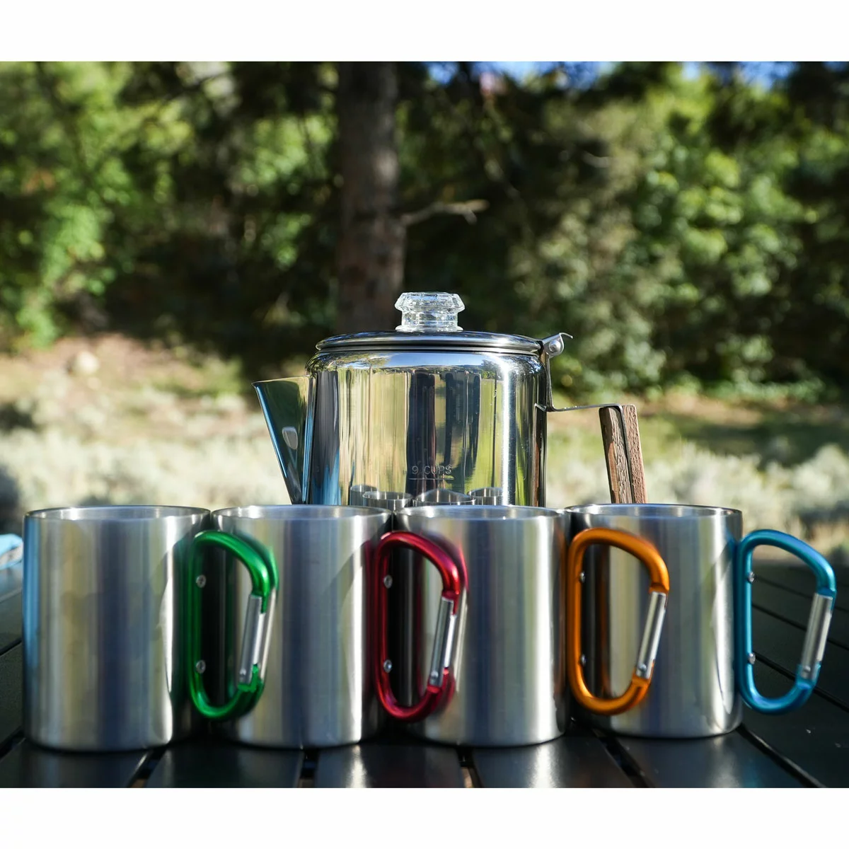 Stainless steel 9-cup Alpine Mountain Gear coffee percolator with glass top knob heating over a campfire next to four carabiner-handle stainless steel mugs on a wooden campsite table.