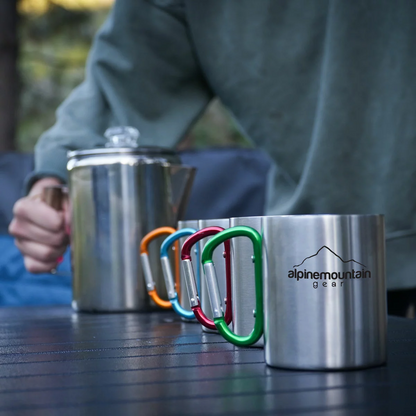 Stainless steel 9-cup Alpine Mountain Gear coffee percolator with glass top knob heating over a campfire next to four carabiner-handle stainless steel mugs on a wooden campsite table.