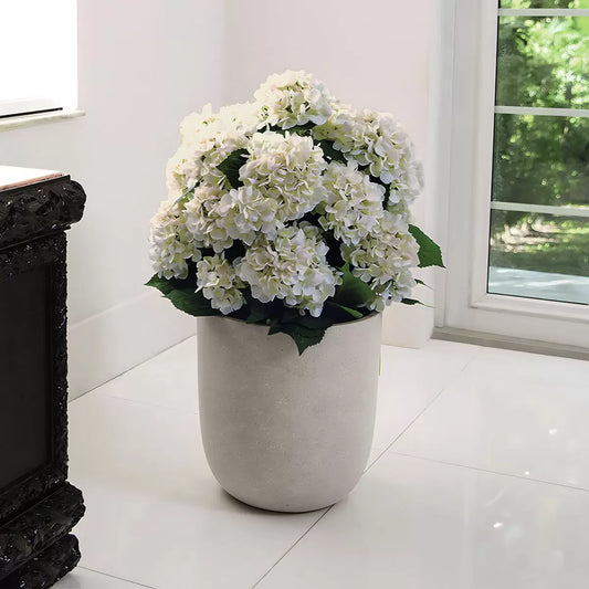 A lush, realistic artificial hydrangea with vibrant blooms in a stylish gray decorative planter sitting on a wooden side table.