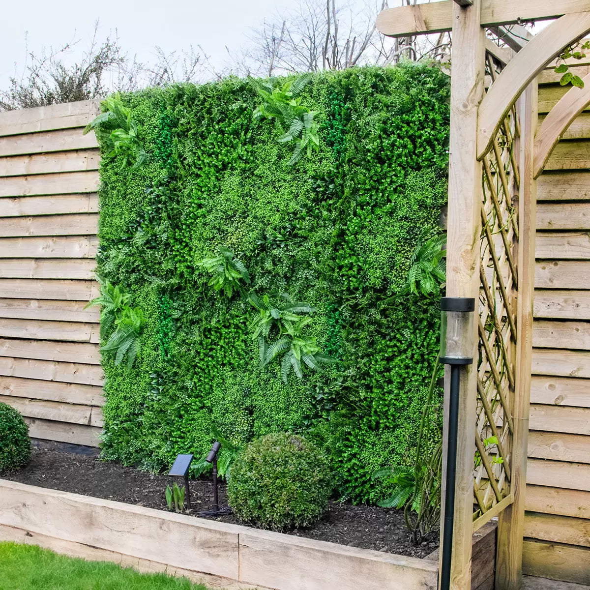 Close-up of deep green artificial mixed foliage panel featuring ivy and fern leaves mounted on an outdoor brick patio wall.