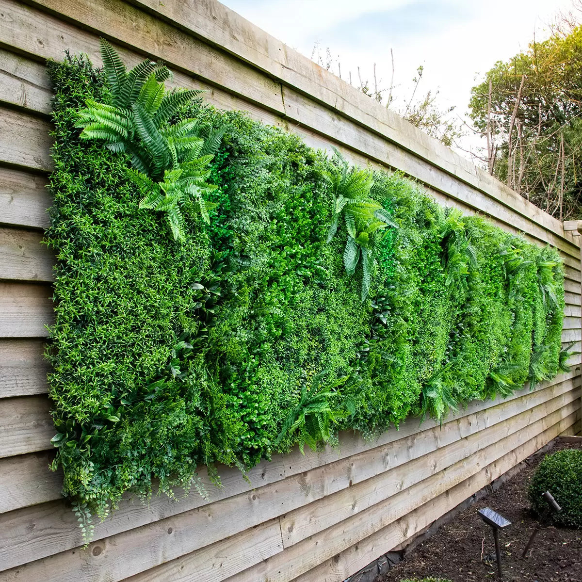 Close-up of deep green artificial mixed foliage panel featuring ivy and fern leaves mounted on an outdoor brick patio wall.