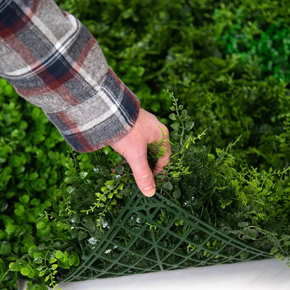 Close-up of deep green artificial mixed foliage panel featuring ivy and fern leaves mounted on an outdoor brick patio wall.