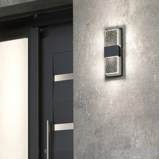 Artika Mazza black wall sconce with a thick vertical glass panel filled with tiny air bubbles, glowing with warm LED light.