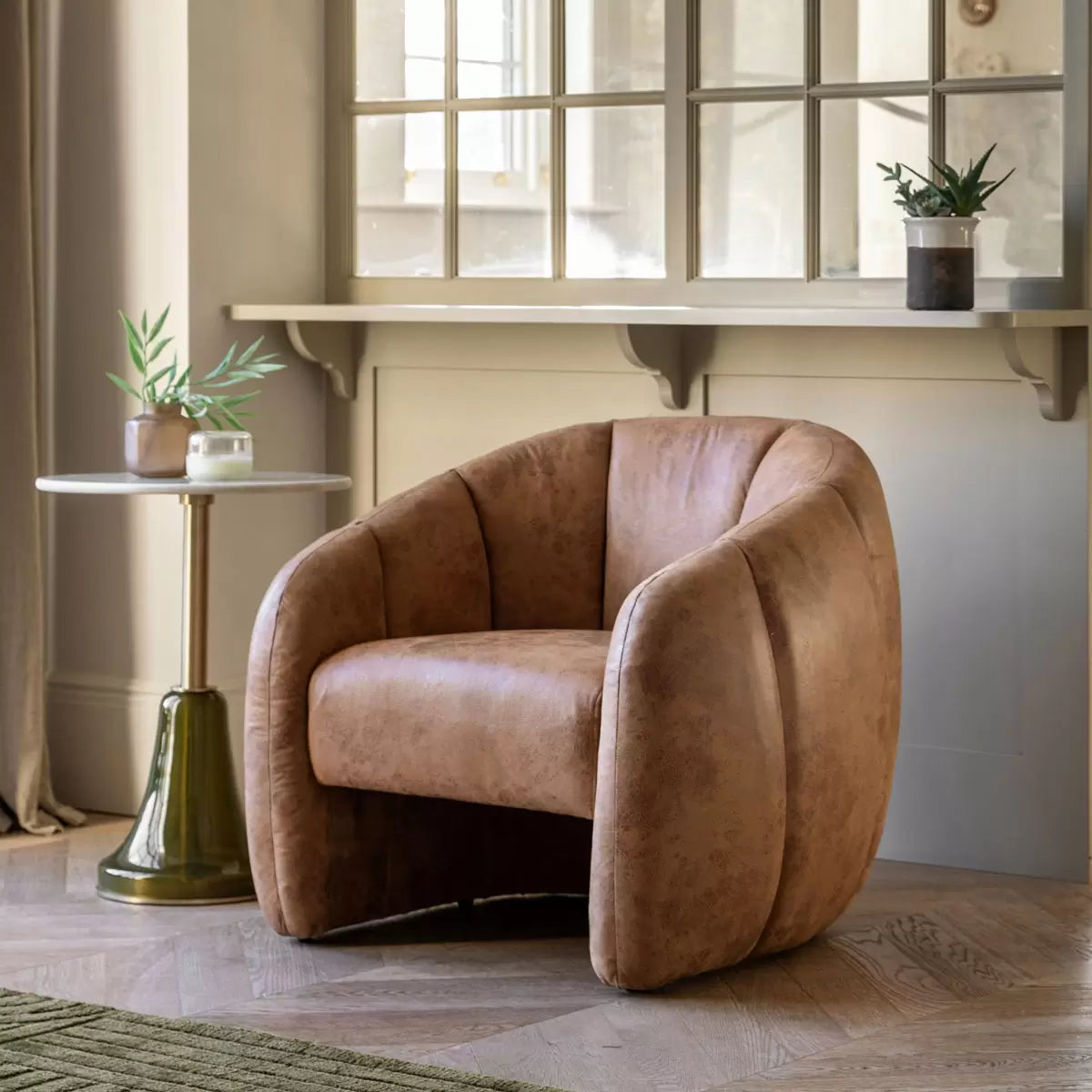 Front three-quarter view of the Gallery Atella Tub Chair in antique tan leather with vertical stitching on the curved backrest, positioned on a rustic wooden floor next to a minimalist brass floor lamp.