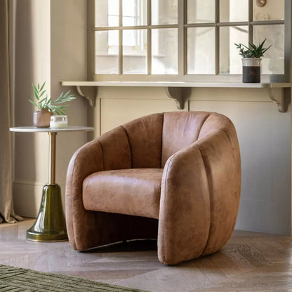 Front three-quarter view of the Gallery Atella Tub Chair in antique tan leather with vertical stitching on the curved backrest, positioned on a rustic wooden floor next to a minimalist brass floor lamp.