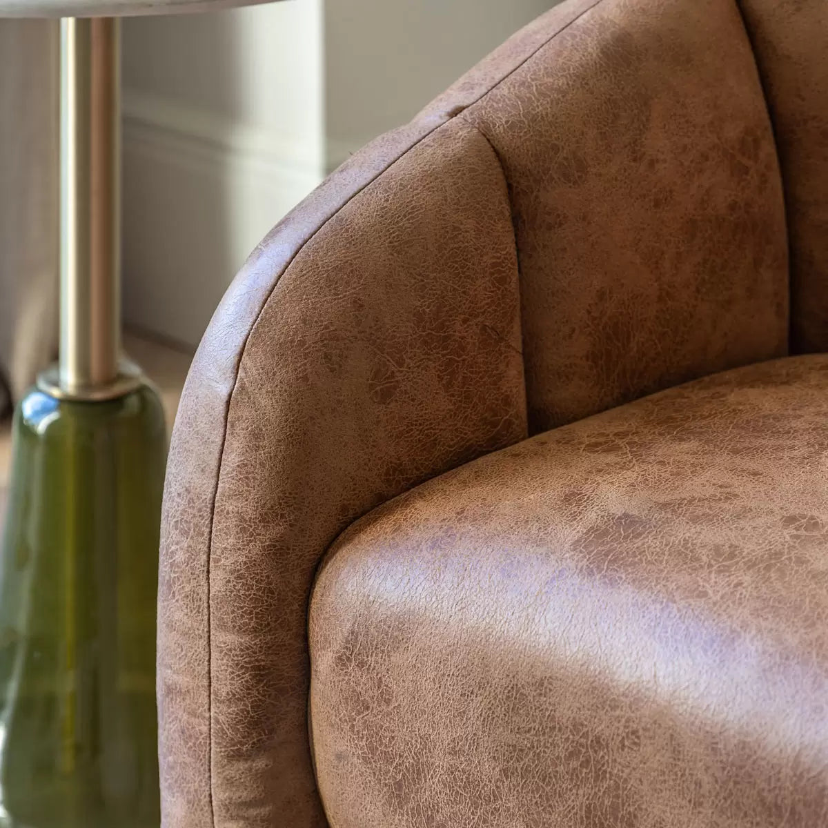 Front three-quarter view of the Gallery Atella Tub Chair in antique tan leather with vertical stitching on the curved backrest, positioned on a rustic wooden floor next to a minimalist brass floor lamp.