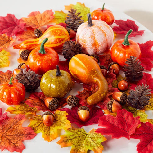 Textured orange, green, and mini artificial pumpkins paired with fabric autumn leaves and pine cones arranged neatly on a wooden table setting.