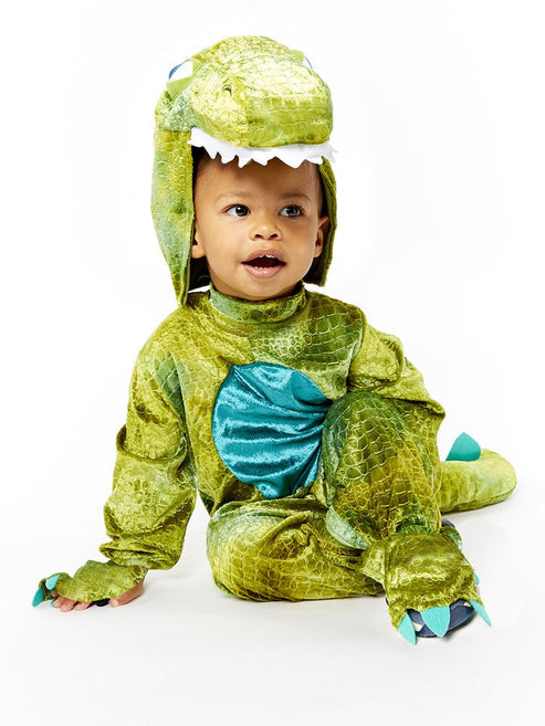 Front view of a toddler wearing a soft green scale-patterned dinosaur jumpsuit with a plush hood and yellow velour belly panel in a bright living room.