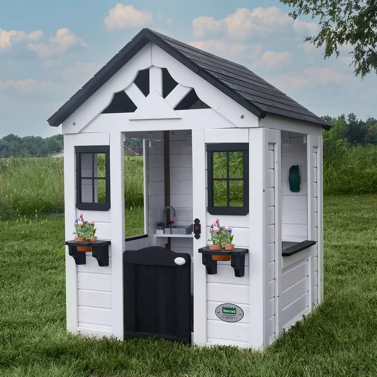 White cedar Backyard Discovery Sweetwater playhouse with black window trim, flowerpot holders, half door, and snack window set up on a green grass garden lawn.