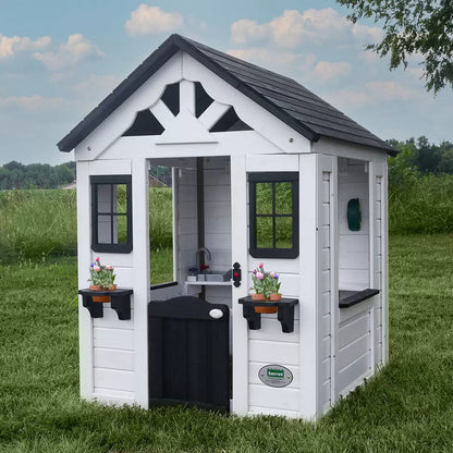 White cedar Backyard Discovery Sweetwater playhouse with black window trim, flowerpot holders, half door, and snack window set up on a green grass garden lawn.