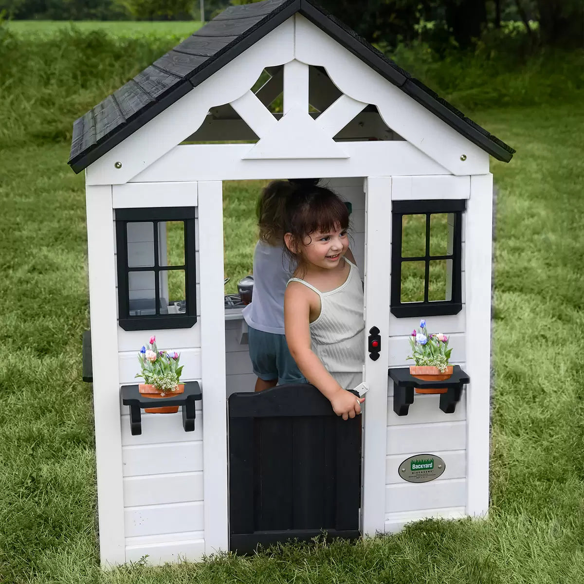 White cedar Backyard Discovery Sweetwater playhouse with black window trim, flowerpot holders, half door, and snack window set up on a green grass garden lawn.