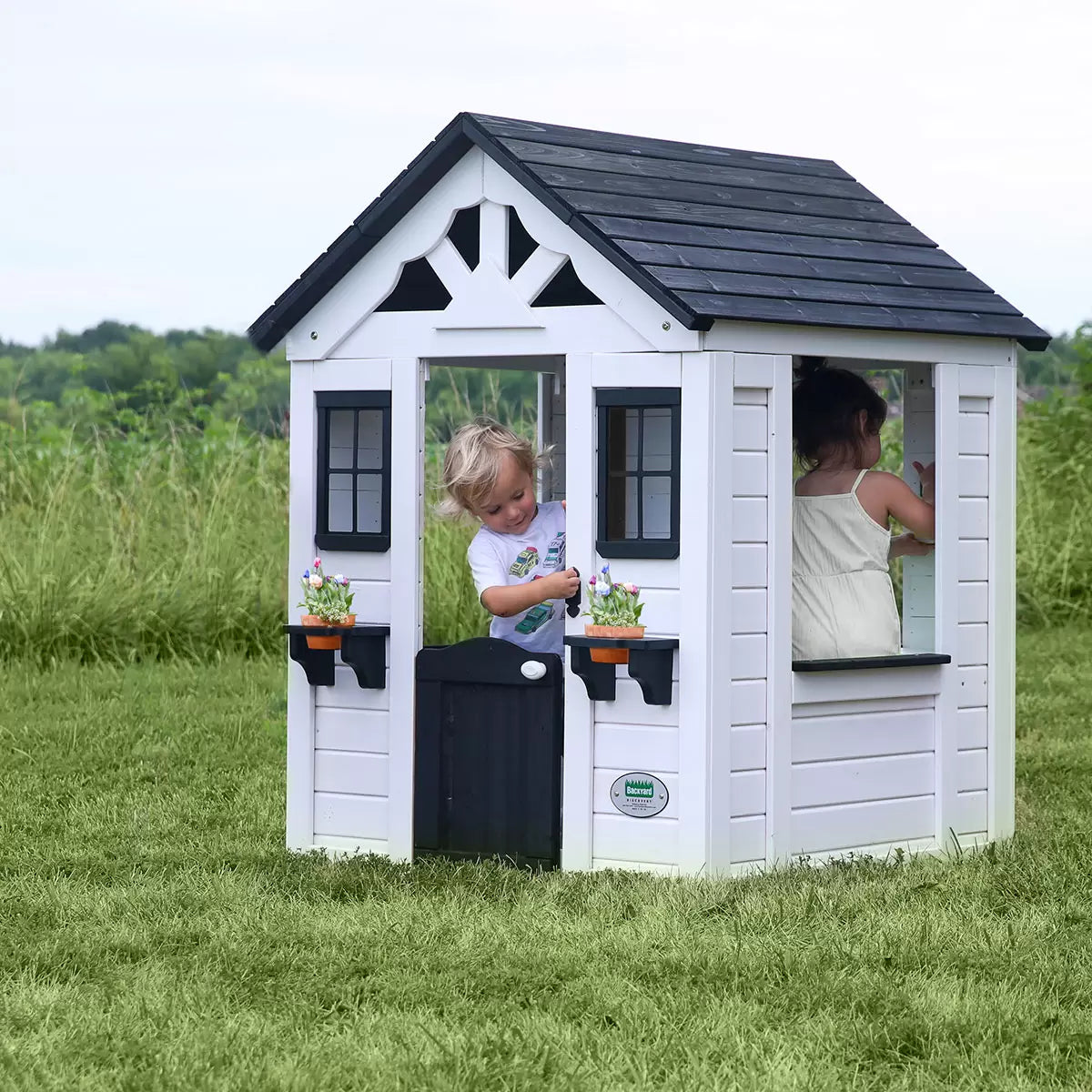 White cedar Backyard Discovery Sweetwater playhouse with black window trim, flowerpot holders, half door, and snack window set up on a green grass garden lawn.