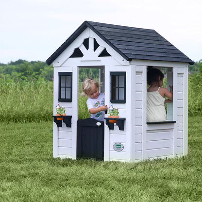 White cedar Backyard Discovery Sweetwater playhouse with black window trim, flowerpot holders, half door, and snack window set up on a green grass garden lawn.