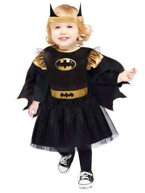 Front view of a toddler wearing the black and yellow officially licensed Batgirl dress with tulle skirt, attached cape, and bat wing headband in a brightly lit play room.