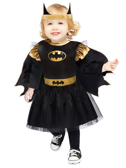 Front view of a toddler wearing the black and yellow officially licensed Batgirl dress with tulle skirt, attached cape, and bat wing headband in a brightly lit play room.