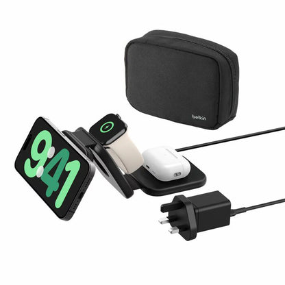 A black foldable Belkin BoostCharge Pro 3-in-1 charging pad laid out flat on a hotel desk, wirelessly charging an iPhone, Apple Watch, and AirPods case.