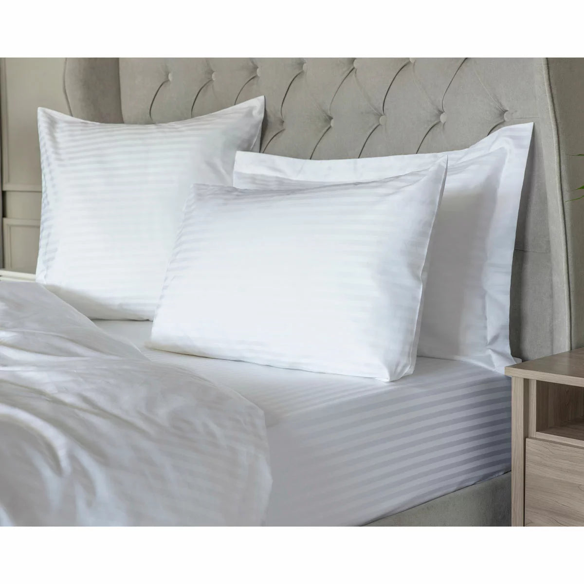 Neat arrangement of the Belledorm 540 thread count white satin stripe duvet cover set with matching pillowcases folded on a minimalist wooden bed frame.